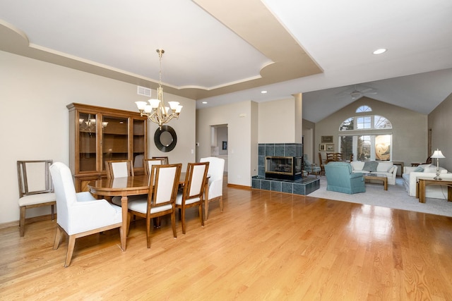 dining space featuring vaulted ceiling, ceiling fan with notable chandelier, a fireplace, light hardwood / wood-style floors, and a raised ceiling