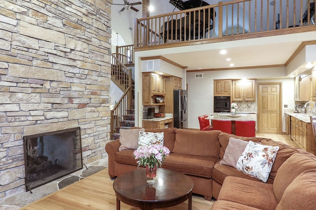 living room with light wood-type flooring, stairway, a stone fireplace, crown molding, and ceiling fan