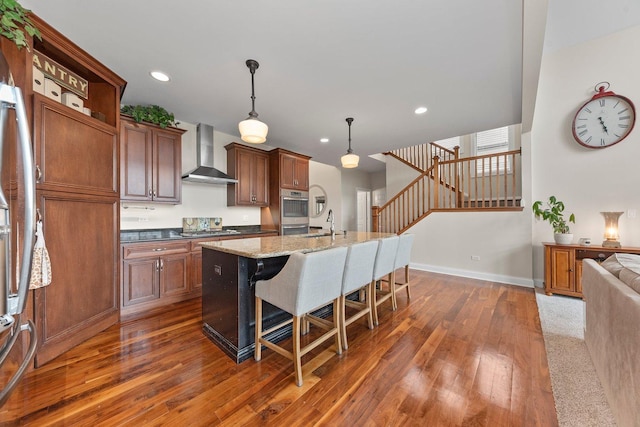 kitchen with a breakfast bar, a kitchen island with sink, wall chimney range hood, decorative light fixtures, and dark hardwood / wood-style floors