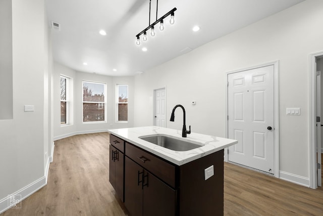 kitchen with a kitchen island with sink, sink, light hardwood / wood-style flooring, light stone counters, and dark brown cabinetry