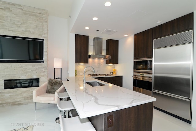 kitchen with wall chimney exhaust hood, tasteful backsplash, built in appliances, kitchen peninsula, and a kitchen bar