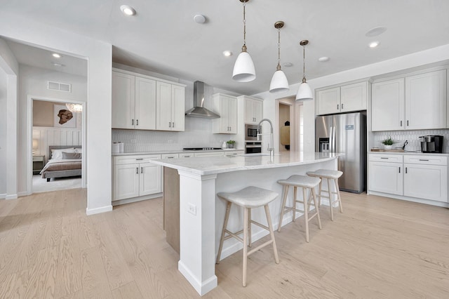 kitchen with light wood-type flooring, tasteful backsplash, wall chimney exhaust hood, stainless steel appliances, and a center island with sink