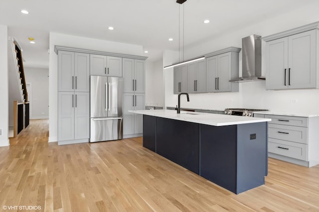 kitchen with pendant lighting, wall chimney exhaust hood, an island with sink, and stainless steel refrigerator