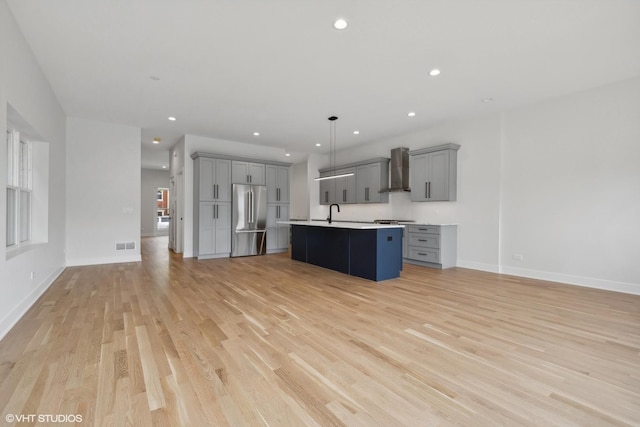 kitchen with gray cabinetry, stainless steel appliances, wall chimney range hood, decorative light fixtures, and a center island with sink