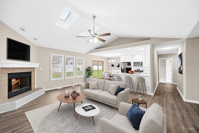 living room featuring a fireplace, vaulted ceiling with skylight, dark wood-type flooring, and ceiling fan with notable chandelier