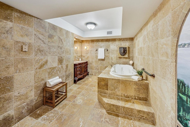 bathroom with a raised ceiling, vanity, tile walls, and a relaxing tiled tub