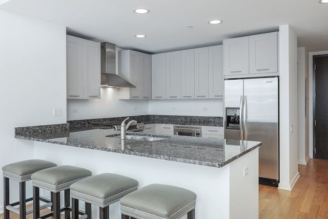 kitchen with kitchen peninsula, appliances with stainless steel finishes, dark stone counters, wall chimney range hood, and light hardwood / wood-style flooring