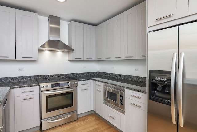 kitchen featuring stainless steel appliances, wall chimney range hood, dark stone countertops, light hardwood / wood-style floors, and white cabinets