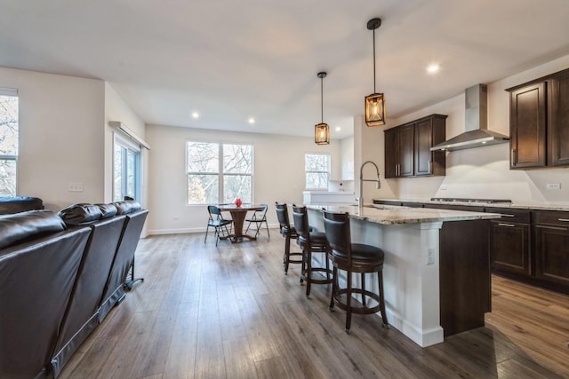 kitchen featuring dark brown cabinetry, wall chimney exhaust hood, dark wood-type flooring, decorative light fixtures, and a center island with sink