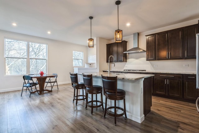 kitchen featuring sink, wall chimney range hood, decorative light fixtures, hardwood / wood-style flooring, and an island with sink