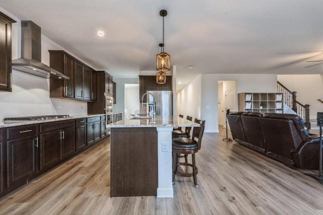 kitchen featuring light wood-type flooring, wall chimney exhaust hood, stainless steel appliances, pendant lighting, and an island with sink