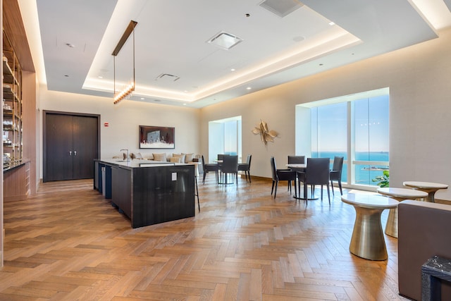 kitchen featuring a raised ceiling, a kitchen island with sink, decorative light fixtures, a water view, and light parquet flooring