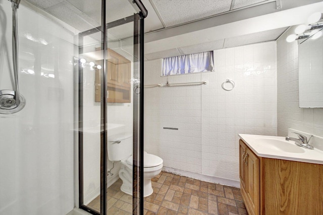 bathroom with toilet, a paneled ceiling, vanity, a shower with shower door, and tile walls