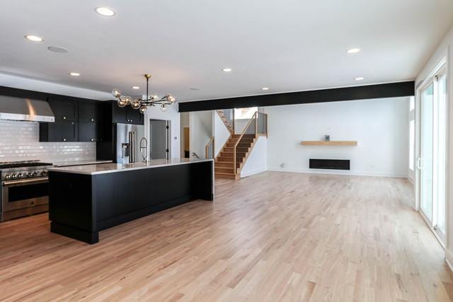 kitchen with wall chimney range hood, light wood-type flooring, an island with sink, decorative light fixtures, and stainless steel appliances