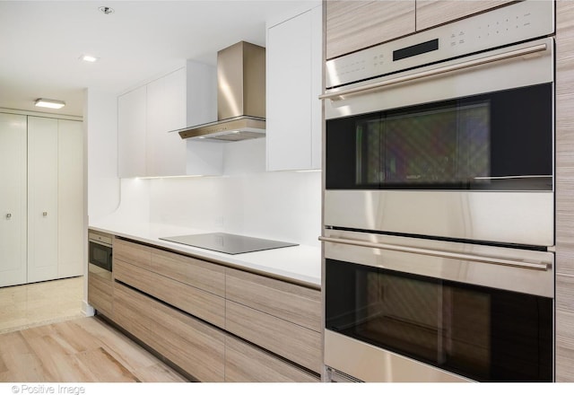 kitchen featuring modern cabinets, black electric cooktop, light countertops, wall chimney range hood, and double oven