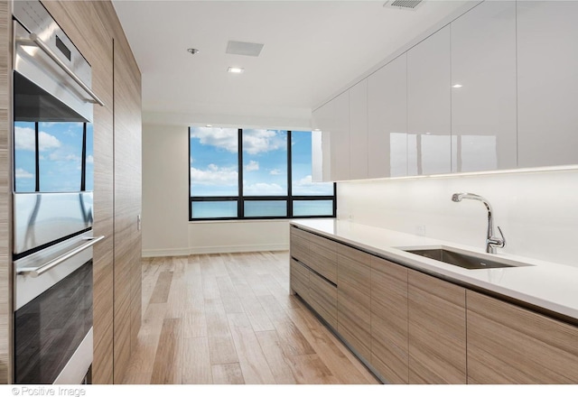 kitchen featuring stainless steel double oven, a sink, light countertops, light wood finished floors, and modern cabinets