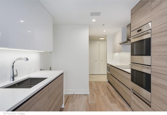 kitchen featuring double oven, a sink, light countertops, wall chimney range hood, and modern cabinets