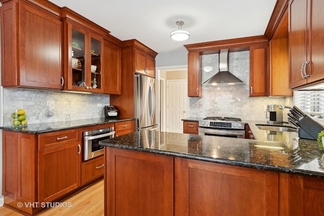 kitchen with dark stone counters, wall chimney exhaust hood, glass insert cabinets, appliances with stainless steel finishes, and a sink