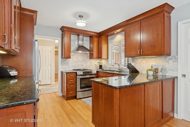 kitchen featuring light wood finished floors, appliances with stainless steel finishes, a sink, a peninsula, and wall chimney exhaust hood