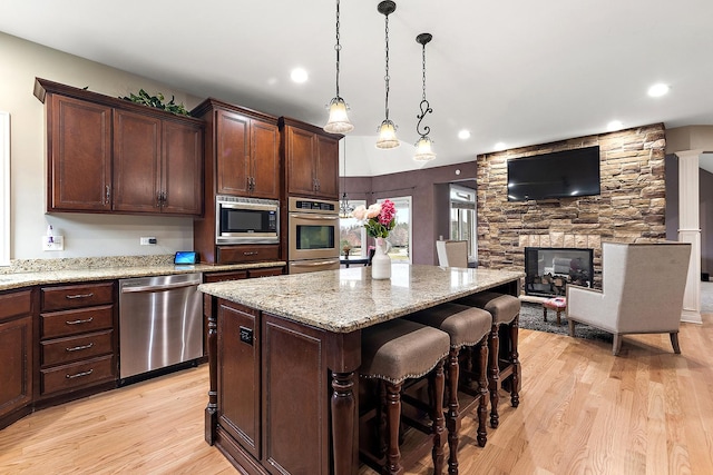 kitchen with appliances with stainless steel finishes, decorative light fixtures, light hardwood / wood-style floors, a kitchen island, and a stone fireplace