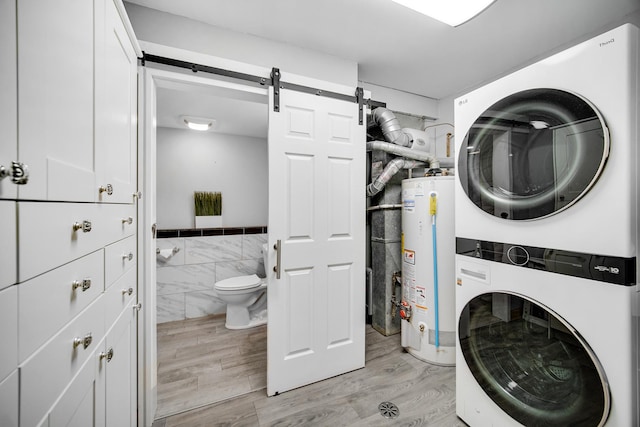 laundry area with light hardwood / wood-style flooring, a barn door, tile walls, water heater, and stacked washer / dryer