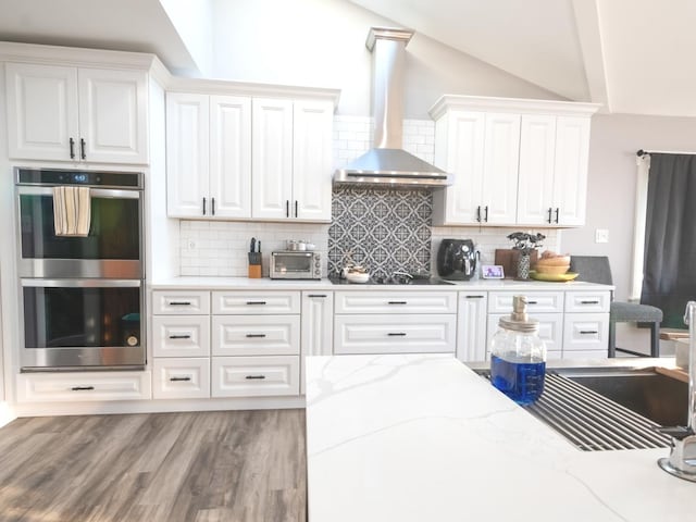 kitchen featuring wall chimney exhaust hood, stainless steel double oven, tasteful backsplash, light hardwood / wood-style floors, and white cabinetry