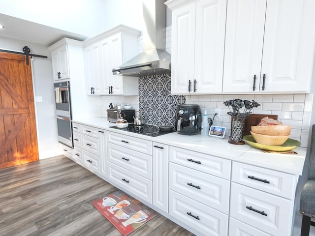 kitchen featuring white cabinetry, wall chimney exhaust hood, a barn door, decorative backsplash, and black electric stovetop