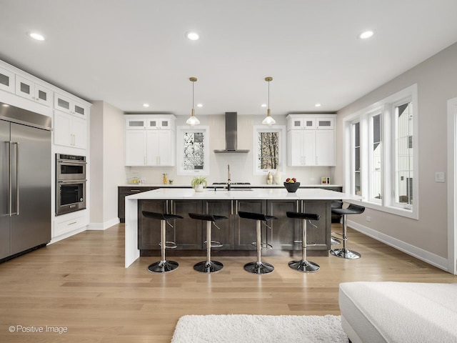 kitchen with white cabinets, a center island with sink, stainless steel appliances, and wall chimney exhaust hood