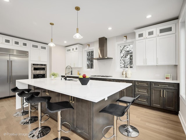 kitchen featuring pendant lighting, white cabinetry, wall chimney range hood, stainless steel appliances, and a center island with sink