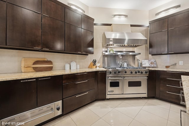 kitchen featuring wall chimney range hood, backsplash, dark brown cabinets, light stone counters, and range with two ovens