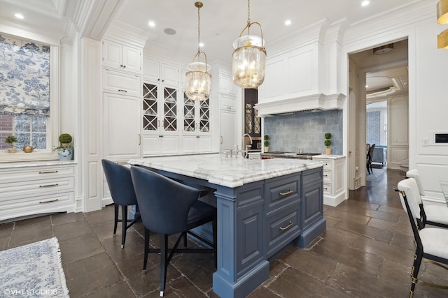 kitchen with a kitchen island with sink, hanging light fixtures, decorative backsplash, light stone counters, and white cabinetry