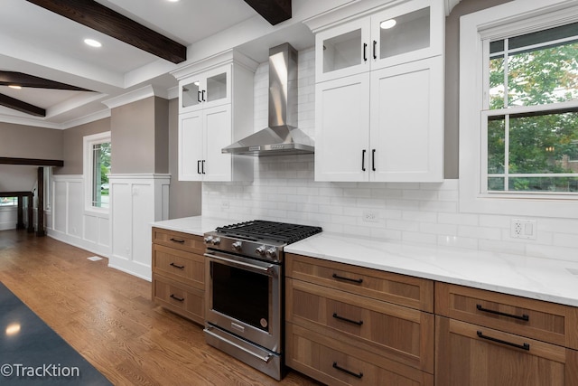 kitchen featuring high end range, white cabinets, wall chimney range hood, beamed ceiling, and light hardwood / wood-style floors