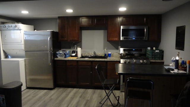 kitchen featuring sink, stainless steel appliances, stacked washer and dryer, dark brown cabinets, and light wood-type flooring