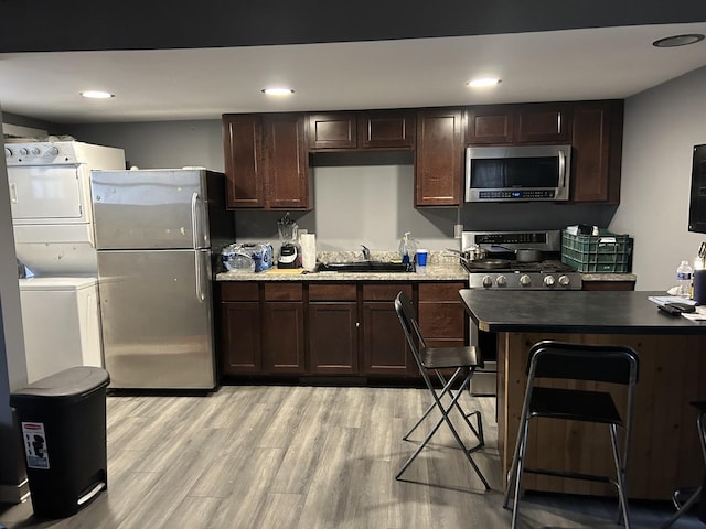 kitchen with dark brown cabinetry, sink, stacked washer / dryer, appliances with stainless steel finishes, and light wood-type flooring