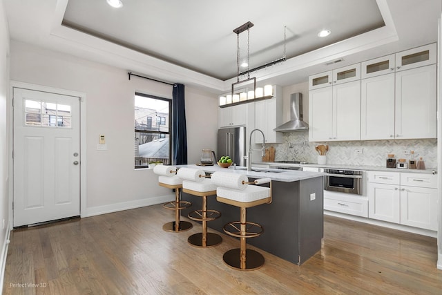 kitchen featuring a tray ceiling, stainless steel appliances, wall chimney range hood, white cabinetry, and an island with sink