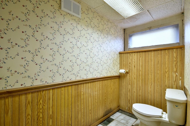 bathroom with toilet, a drop ceiling, and wood walls