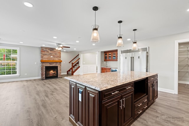 kitchen with a stone fireplace, built in refrigerator, dark brown cabinets, light wood-type flooring, and pendant lighting