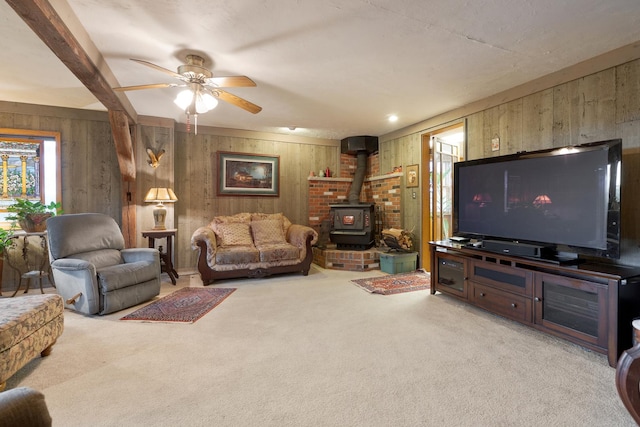 carpeted living room with beamed ceiling, a wood stove, ceiling fan, and wood walls