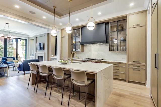 kitchen with custom exhaust hood, hanging light fixtures, a tray ceiling, a kitchen island with sink, and backsplash