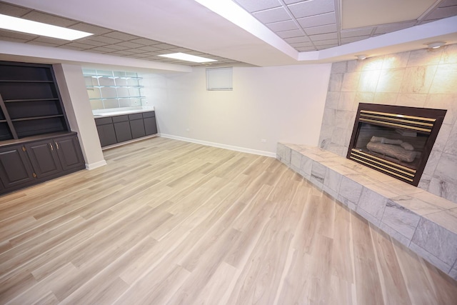 basement with a paneled ceiling, built in shelves, a tile fireplace, and light hardwood / wood-style flooring