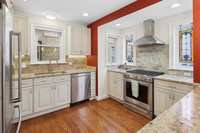 kitchen with a sink, wood finished floors, stainless steel appliances, wall chimney exhaust hood, and light stone countertops