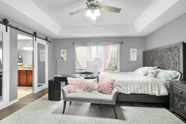 bedroom featuring a raised ceiling, ensuite bath, ceiling fan, a barn door, and dark hardwood / wood-style floors