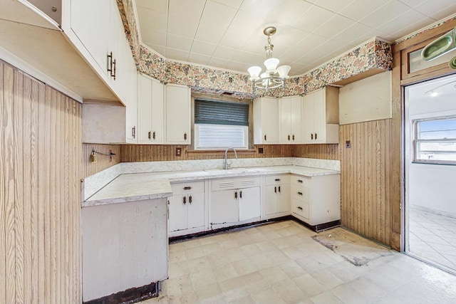 kitchen with wooden walls, sink, an inviting chandelier, white cabinetry, and hanging light fixtures