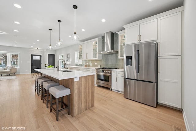 kitchen with a sink, white cabinetry, appliances with stainless steel finishes, wall chimney range hood, and light wood-type flooring