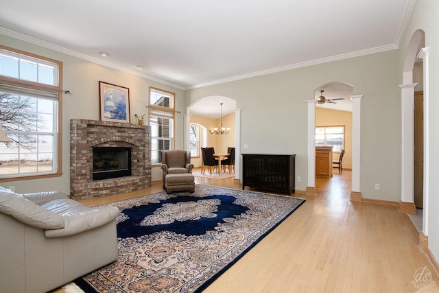 living room featuring crown molding, a brick fireplace, light hardwood / wood-style floors, ceiling fan with notable chandelier, and decorative columns