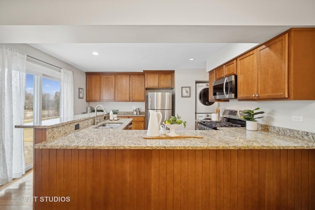 kitchen featuring sink, stainless steel appliances, stacked washer and clothes dryer, light stone countertops, and kitchen peninsula