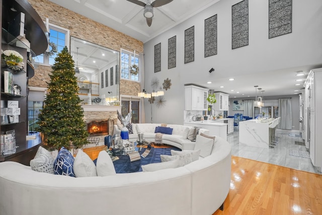 living room with light wood-type flooring, a towering ceiling, coffered ceiling, ceiling fan, and a fireplace