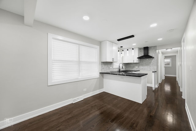 kitchen featuring dark countertops, decorative light fixtures, a peninsula, wall chimney range hood, and white cabinetry