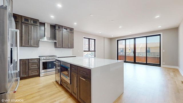 kitchen featuring wall chimney range hood, light hardwood / wood-style flooring, dark brown cabinets, a kitchen island, and appliances with stainless steel finishes