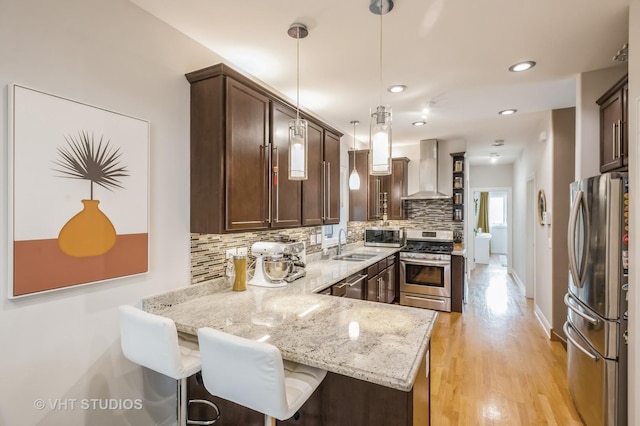 kitchen with light stone countertops, wall chimney range hood, kitchen peninsula, decorative light fixtures, and appliances with stainless steel finishes